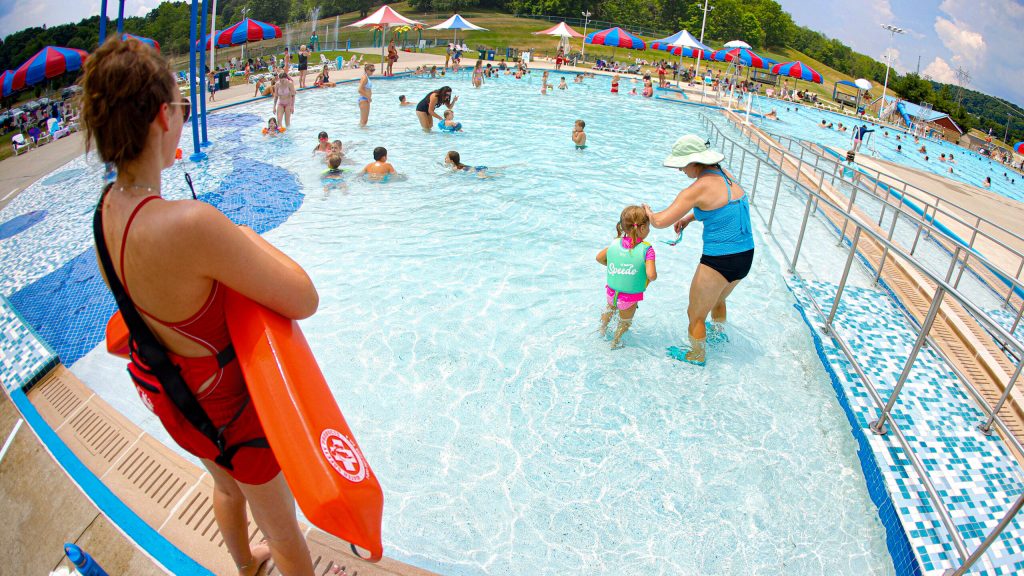 POV lifeguard view of busy outdoor pool with red rescue tube swimmers under colorful umbrellas Wallpaper