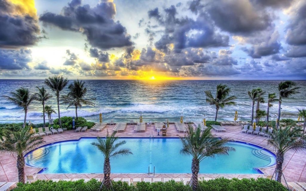 Sunset over resort pool and palm trees with ocean waves for tranquil coastal wallpaper background