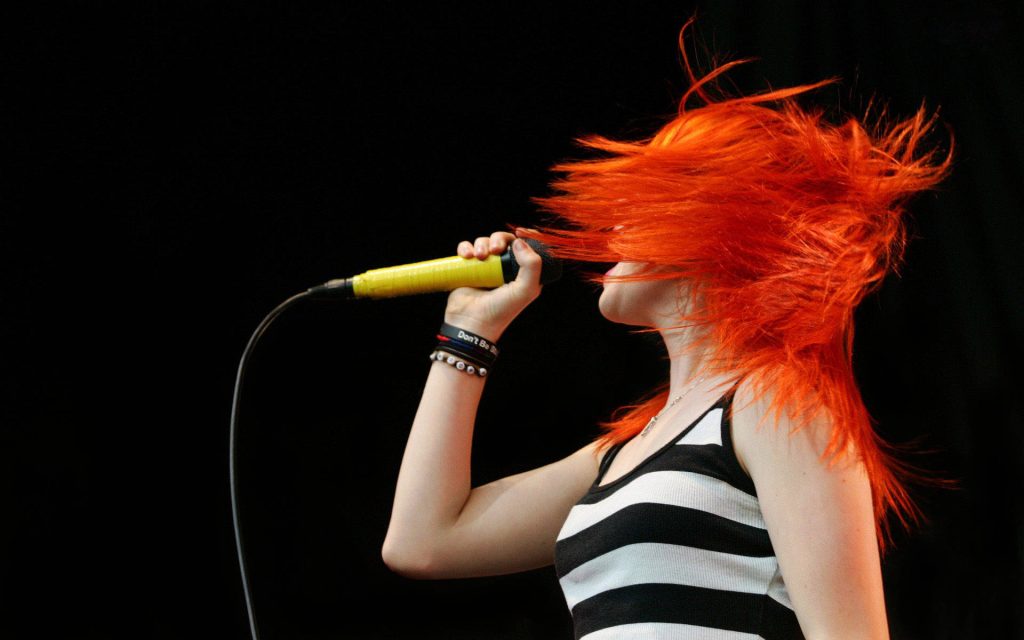 Paramore Hayley Williams mid-performance with orange hair singing into yellow mic on black background Wallpaper