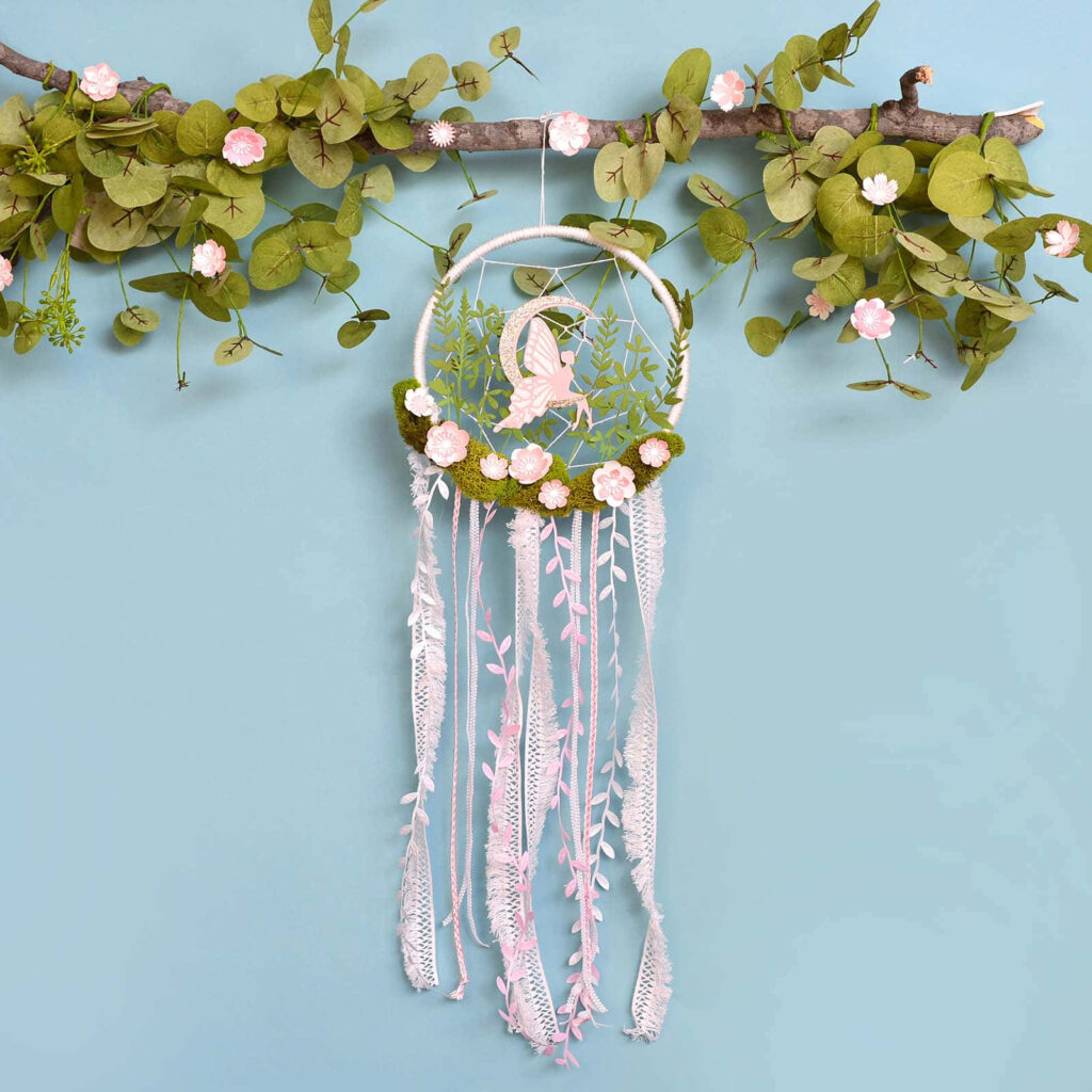 Dream catcher with pastel pink flowers and lace ribbons hanging from leafy branch on blue background Wallpaper