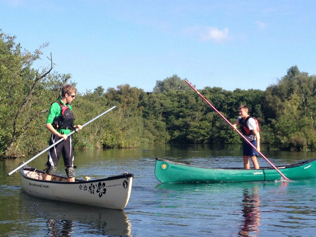 Two people canoeing on calm water surrounded by green trees and blue sky wallpaper background