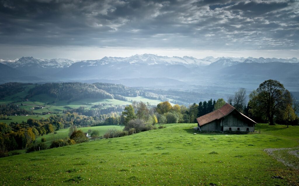 Countryside lodge on green plains with rolling hills and mountains under dramatic cloudy sky Wallpaper