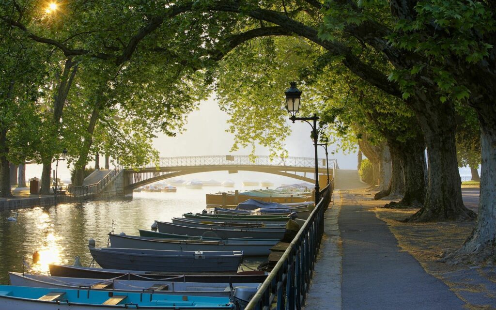 Peaceful French riverside with moored boats shaded by trees and a charming bridge background Wallpaper