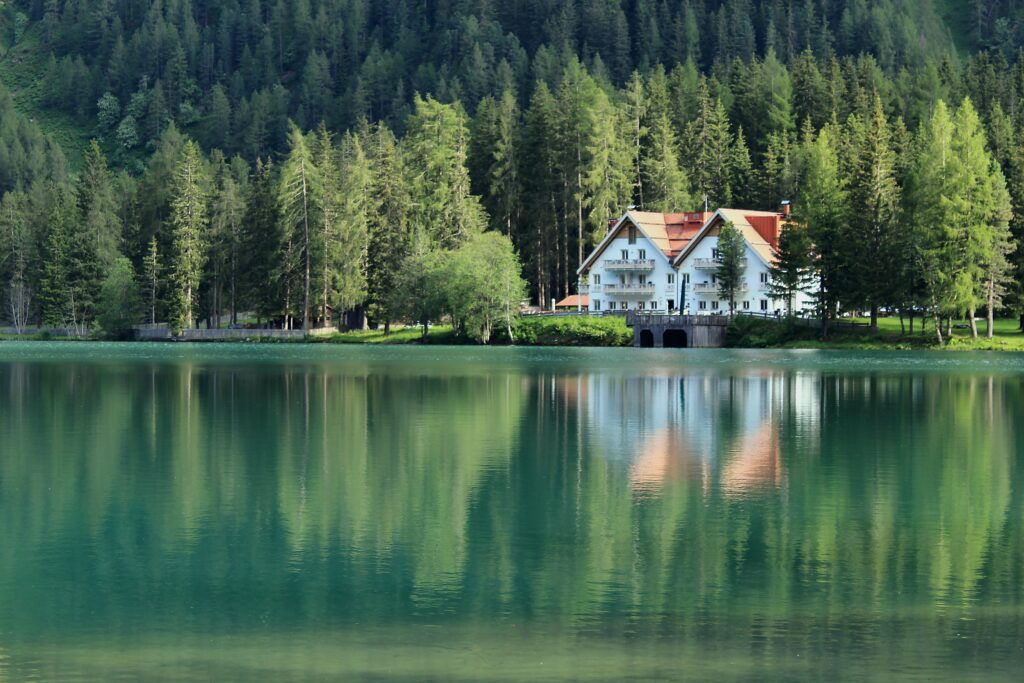 Charming white lakeside building reflected on calm turquoise water with tall evergreens background Wallpaper