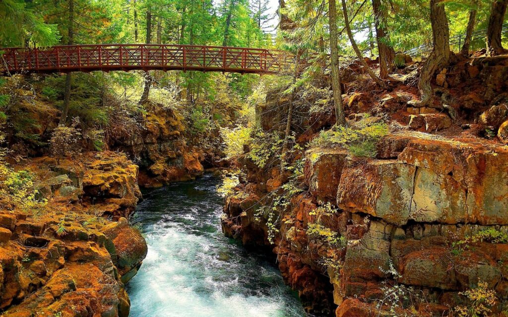 Peaceful river flowing between rugged rocks and lush green trees beneath wooden bridge wallpaper