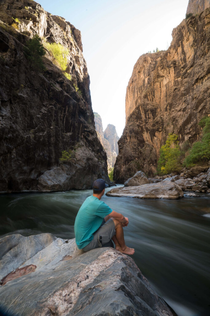 Man sitting on riverside rock gazing at sunlit towering cliffs of Black Canyon landscape wallpaper