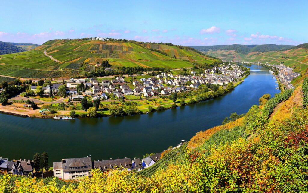 Top view of peaceful riverside village with lush green grasslands and rolling hills under blue sky Wallpaper