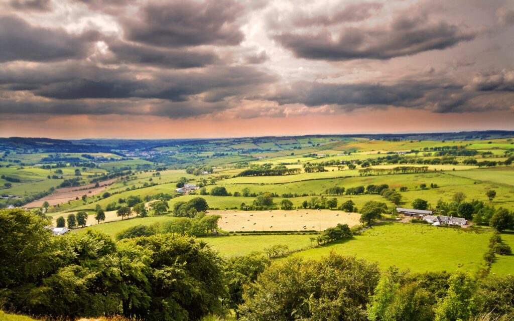 Vast rural plain with scattered lodges and trees under dramatic cloud-filled sky at sunset Wallpaper