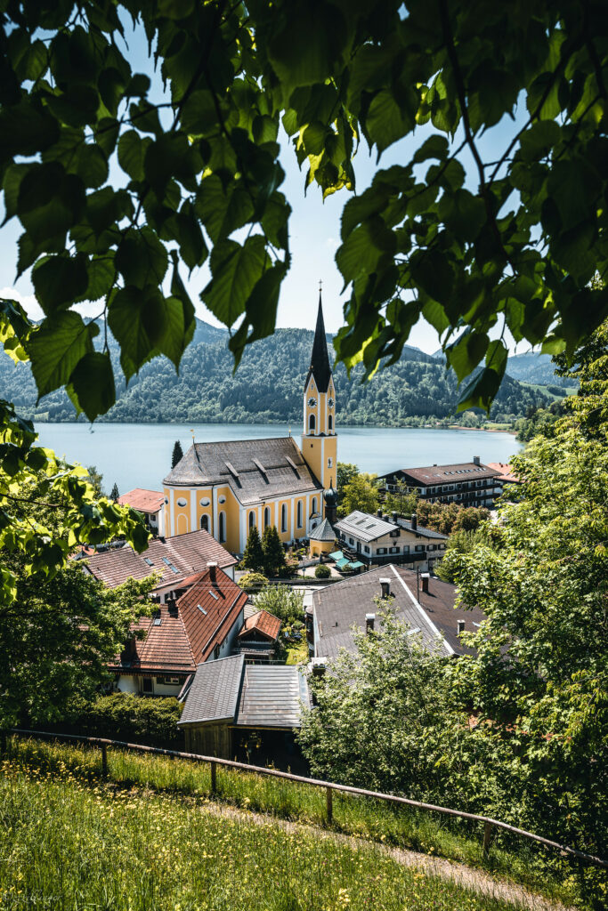 Charming village chapel with tall steeple by lake and forested mountains framed by green leaves Wallpaper
