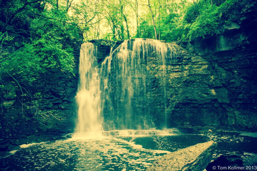 Gentle waterfall over layered rocks with lush green trees and vibrant foliage in soft sunlight Wallpaper