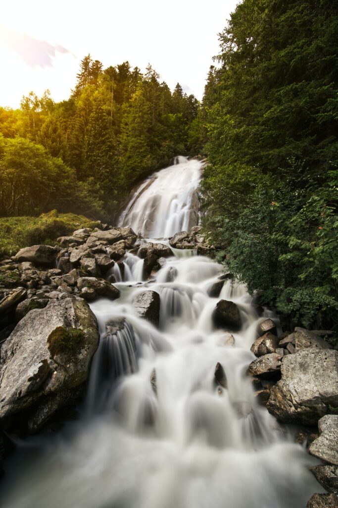 Serene waterfall flowing over rocks with lush green trees and a gentle stream background Wallpaper