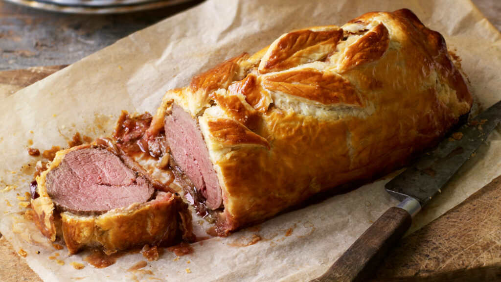 Golden flaky Beef Wellington on parchment paper with tenderloin center and nearby knife ready Wallpaper