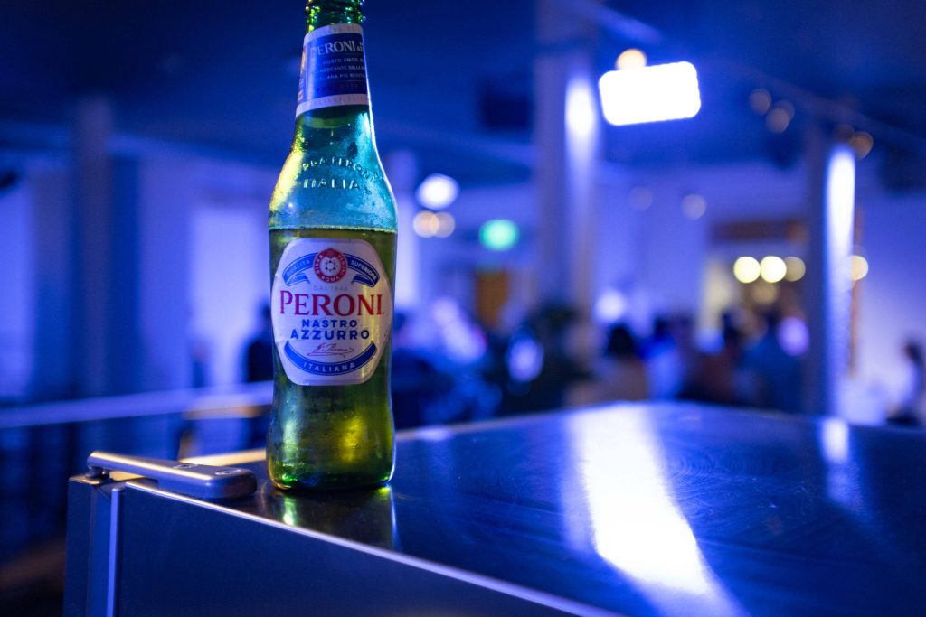 Chilled Peroni Nastro Azzurro bottle on reflective surface with vibrant blue nightlife background Wallpaper