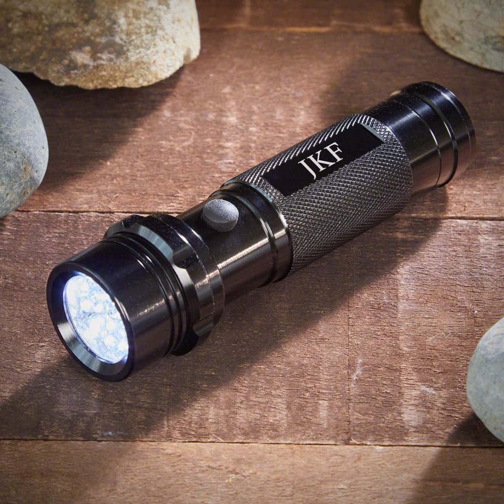 Black LED flashlight engraved JKF glowing on rustic wood and pebbles tech wallpaper background