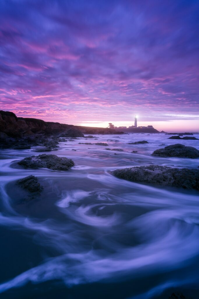 Pescadero lighthouse glowing at night with waves on rocky coast under purple pink twilight sky Wallpaper