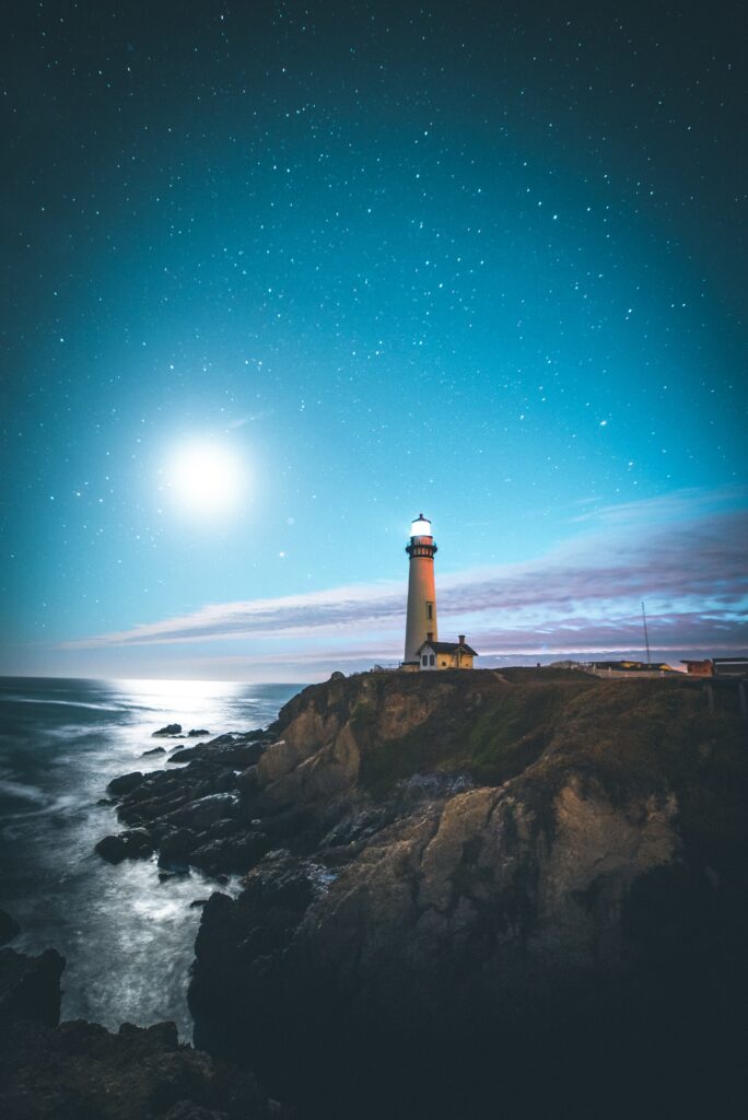 Pescadero lighthouse on rugged US coast illuminated under starry sky with calm ocean background Wallpaper