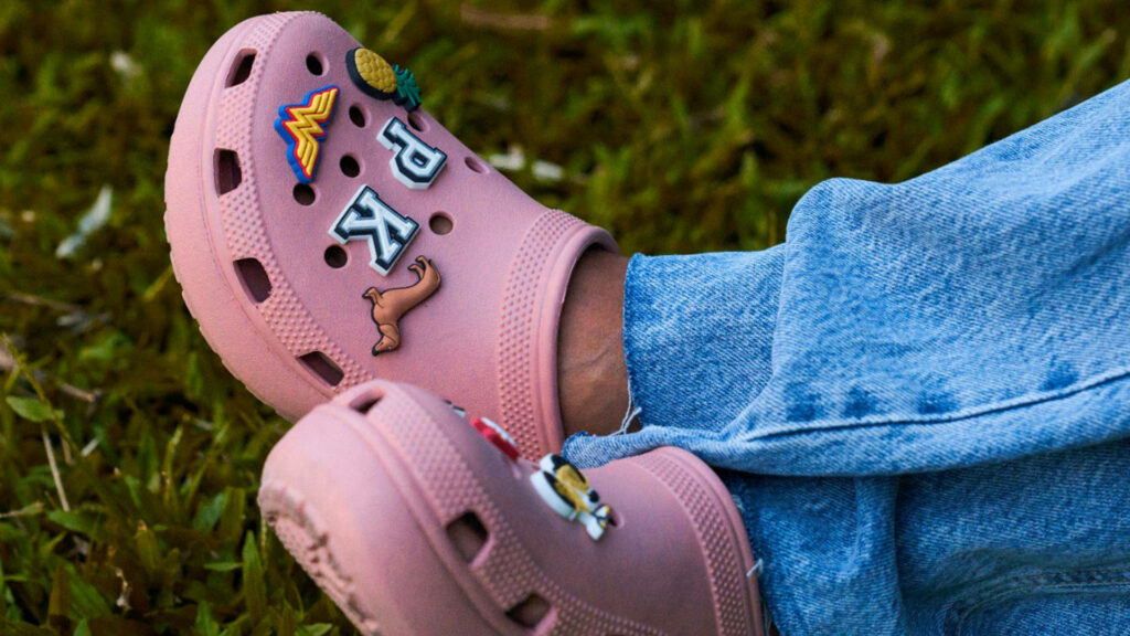 Close-up of pink Crocs with colorful Jibbitz charms on grass worn with light blue denim jeans Wallpaper
