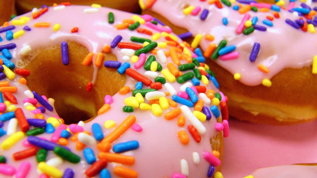 Sugar-Coated Delights: Vibrant Sprinkle-Topped Donut Temptation on Pink Bliss Wallpaper