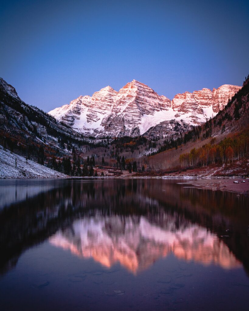 Snow-capped mountains at pink twilight reflected in a clear lake surrounded by autumn trees Wallpaper