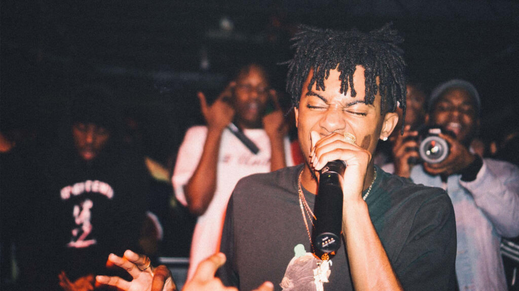 Playboi Carti live concert with dreadlocks rapping moody stage lighting grainy hip hop wallpaper