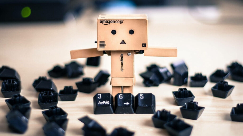 Cardboard Amazon Boxman figure on desk holding keyboard keys 3 6 5 with scattered keys background Wallpaper