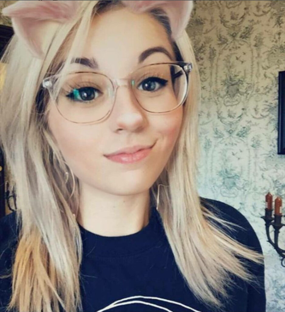 Playful portrait of Amy Nelson wearing glasses with cat filter ears and vibrant eyes on soft wallpaper background