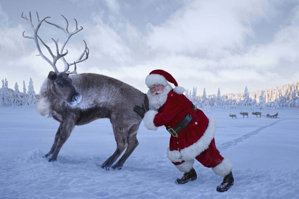 Santa Playfully Pushes Reindeer in Humorous Snowy Landscape Wallpaper