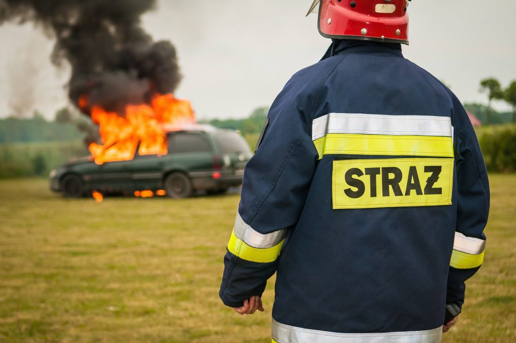 Polish firefighter in STRAZ gear watches blazing car fire with thick black smoke in open field Wallpaper