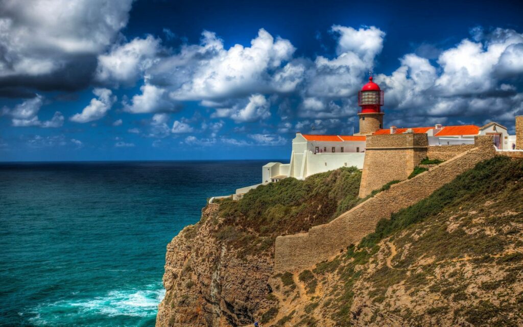 Lighthouse on rugged Portugal cliffs with red roofs white walls overlooking blue Atlantic Ocean Wallpaper
