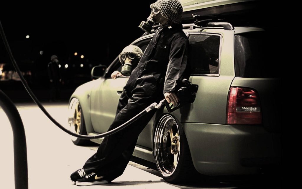 Two figures in gas masks fueling stanced station wagon at dystopian post-apocalyptic gas station Wallpaper