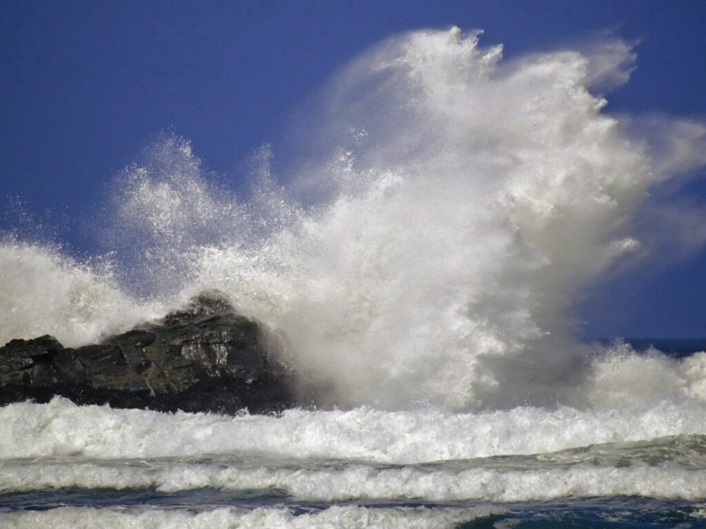 Powerful ocean wave crashing against dark rocks with white sea spray under vivid blue sky wallpaper