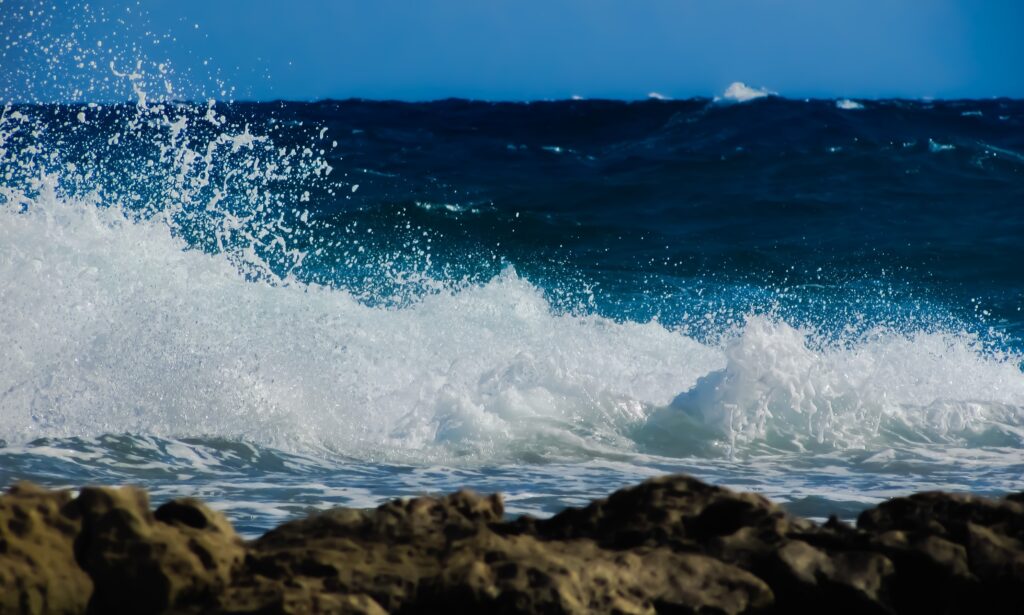 Powerful ocean wave crashing against rocks with white foam spray and deep blue sea tones wallpaper
