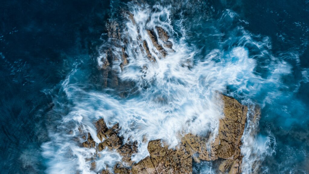 Aerial view of powerful ocean waves crashing on rugged rocks with swirling white sea foam Wallpaper