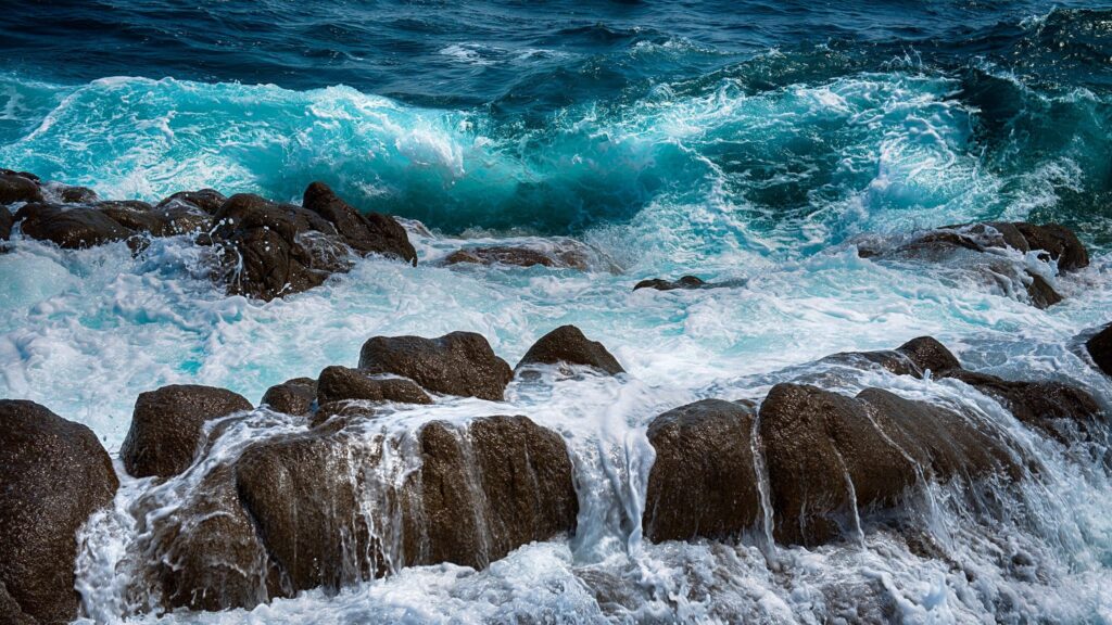 Powerful ocean waves crashing on rugged rocks with white sea spray and vivid blue water background Wallpaper