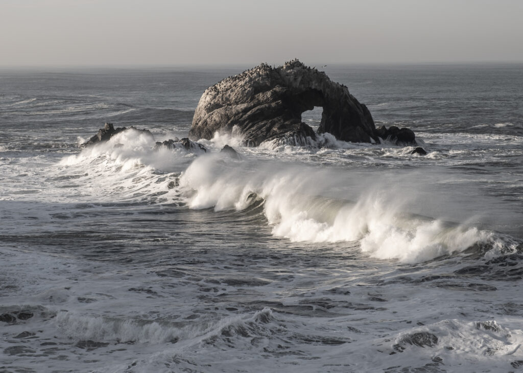 Ocean waves crash rugged rock sending white foam into air under hazy sky dynamic sea nature Wallpaper