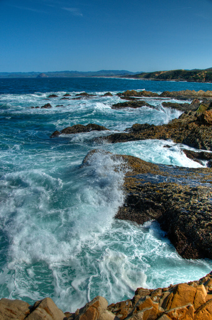 Powerful ocean waves crash on rugged rocks at sunlit shore with vivid blue sea and clear sky Wallpaper
