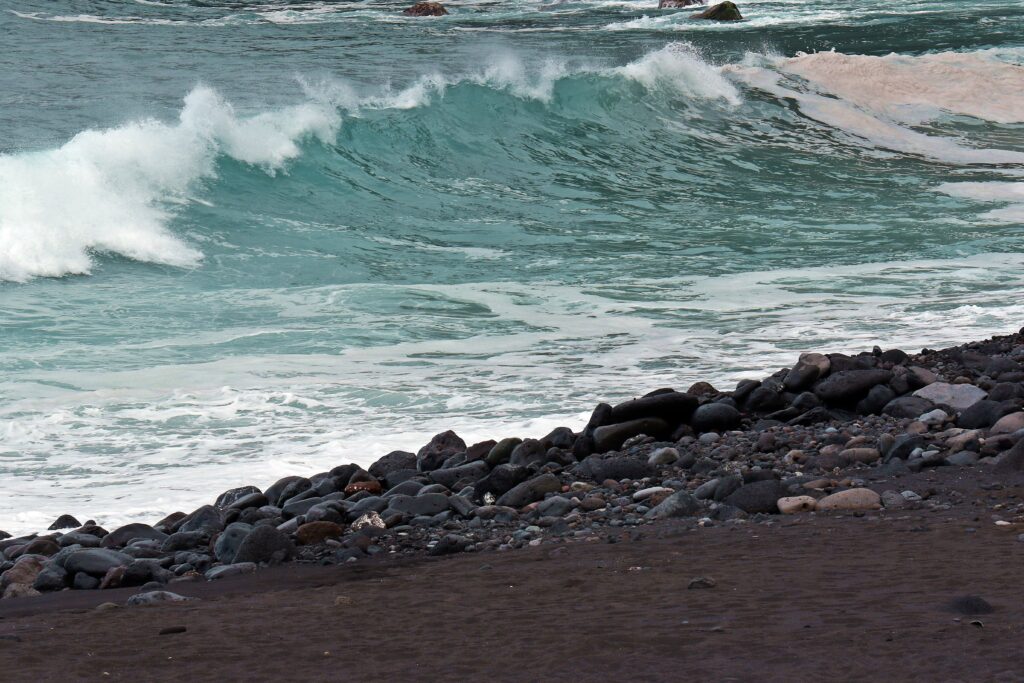 Turquoise Atlantic Ocean waves crash on dark sand rocky beach creating dramatic coastal scene Wallpaper
