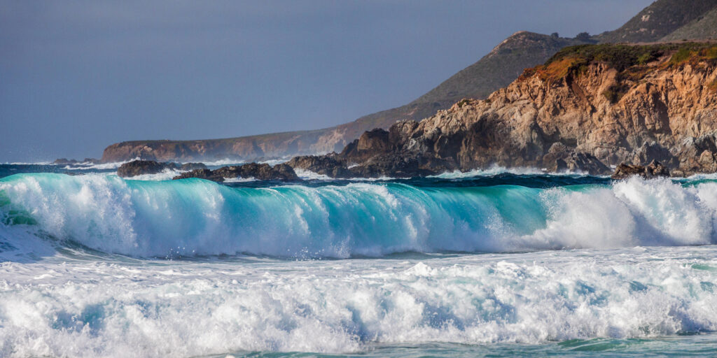 Turquoise waves crash on rugged rocky coast under cloudy sky with sea spray in vibrant seascape Wallpaper