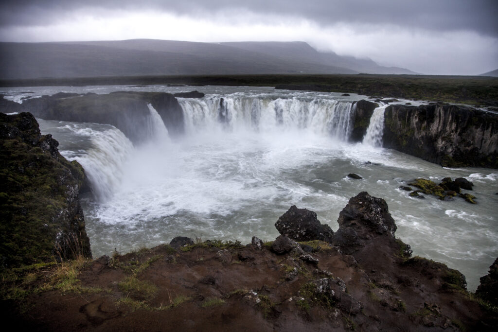 Powerful waterfall cascading over rugged stones with mist and dramatic cliffs under overcast skies Wallpaper