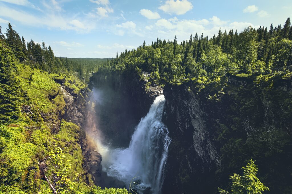 Powerful waterfall cascading over rocks surrounded by lush green forest under blue sky wallpaper