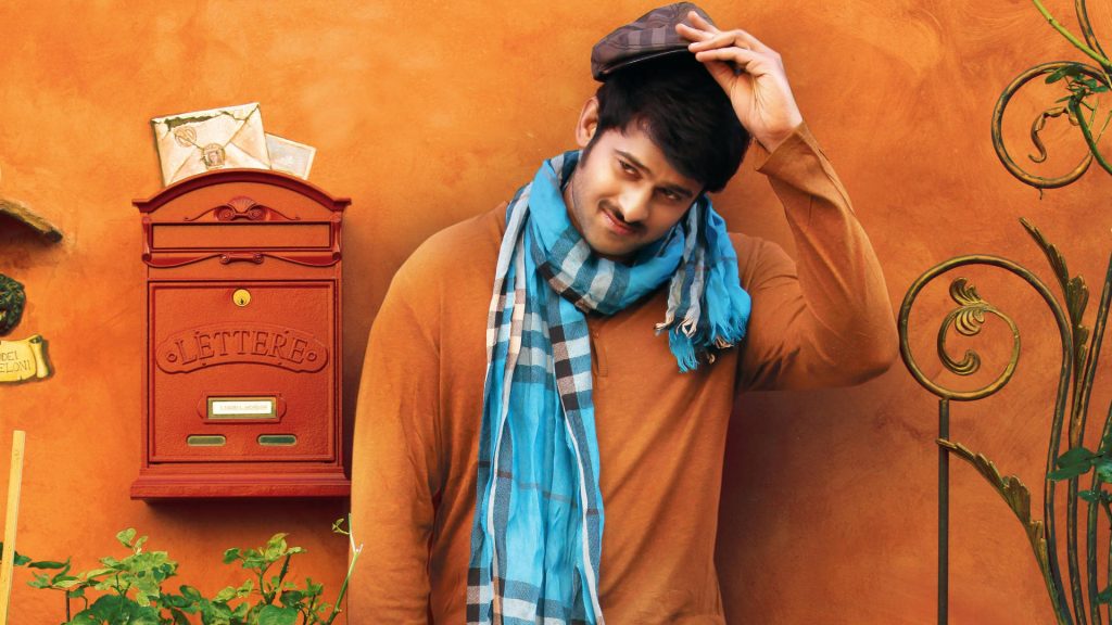 Indian actor Prabhas in brown shirt and blue scarf by vintage red mailbox warm orange backdrop Wallpaper