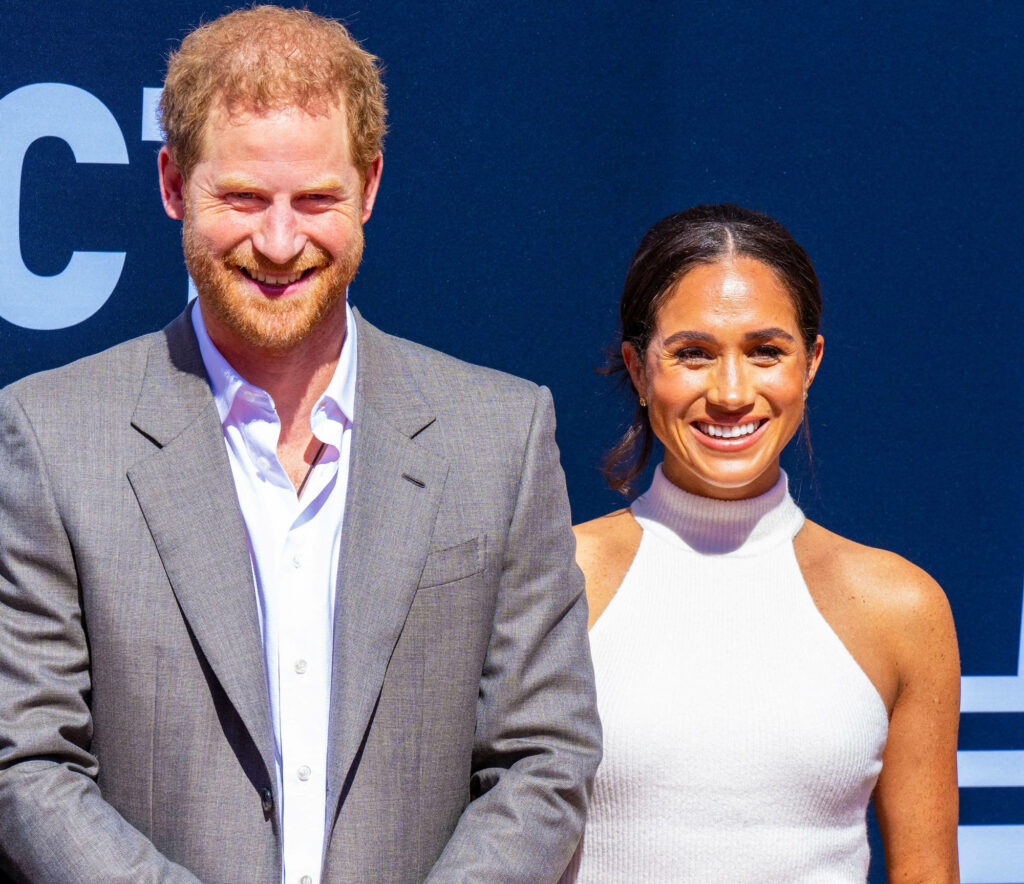 Prince Harry in gray suit and Meghan Markle in white dress smiling against blue backdrop wallpaper