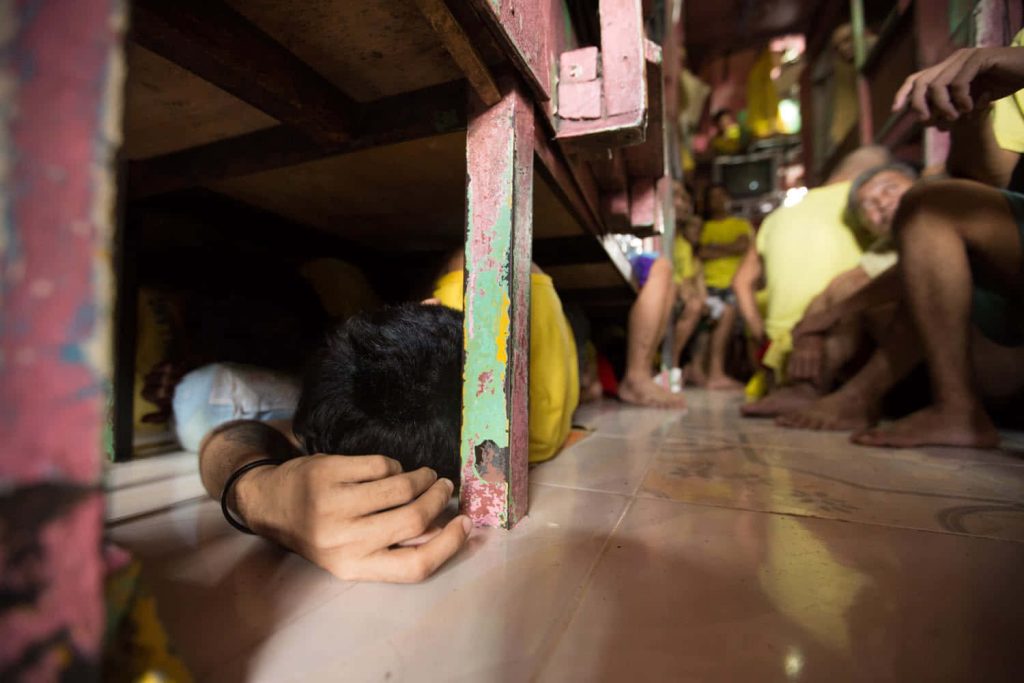 Inmate in yellow uniform resting under bunk bed in crowded dim prison cell showing confinement Wallpaper