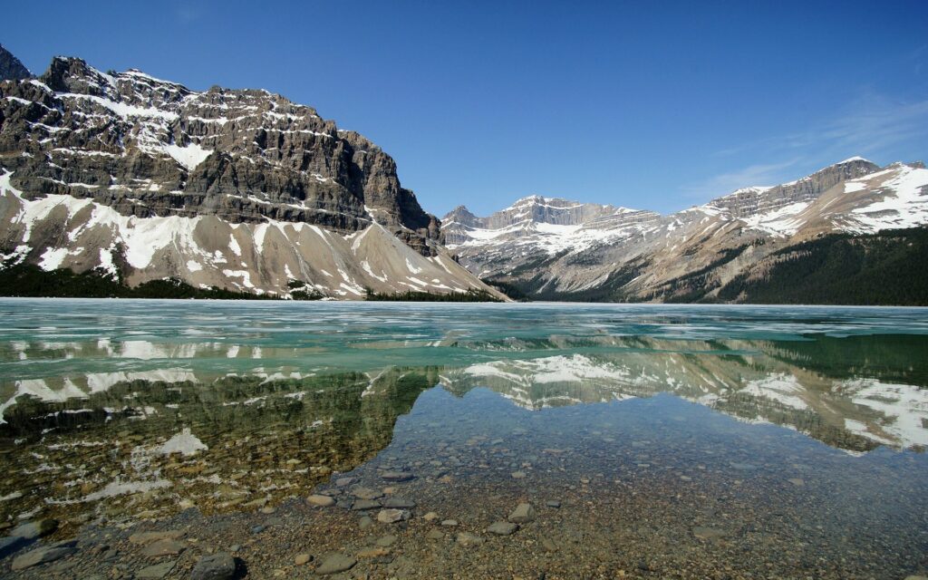 Crystal-clear lake with rocky bottom reflecting snow-capped mountains under blue sky wallpaper