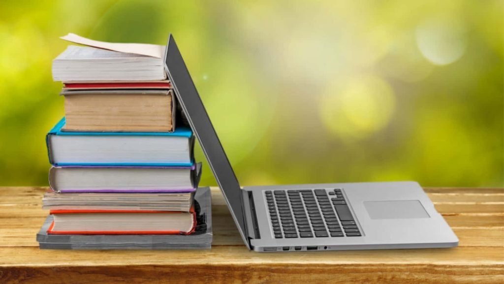 Silver laptop next to tall stack of books on rustic table with bright green natural background Wallpaper