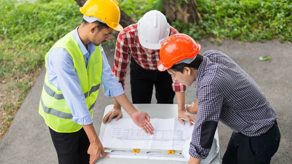 Three engineers in hard hats reviewing architectural blueprints outdoors construction teamwork wallpaper