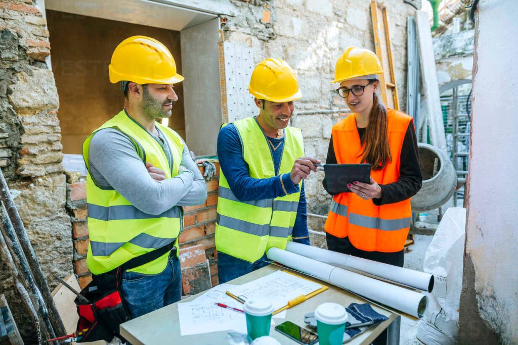 Three construction workers in safety gear reviewing blueprints at active renovation site wallpaper background