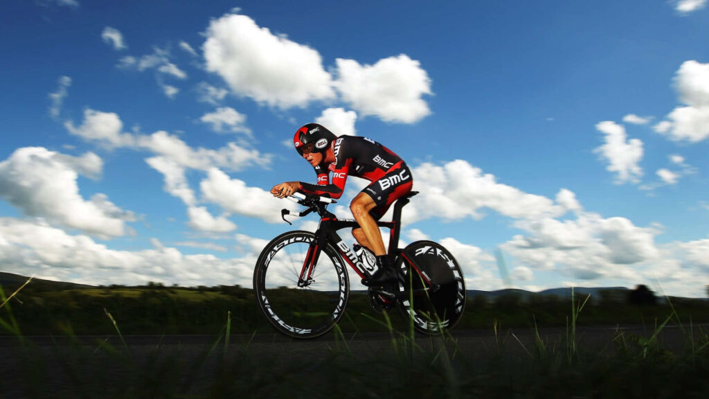 Professional cyclist in BMC gear racing on open road beneath vivid blue sky with white clouds Wallpaper