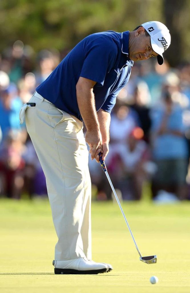 Golfer in navy polo and khaki pants putting on sunny green with blurred spectators behind Wallpaper