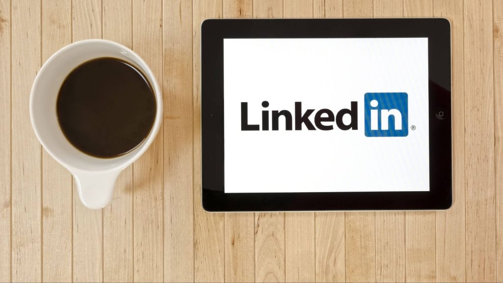 Flat lay of tablet showing LinkedIn logo next to black coffee on light wood desk workspace background Wallpaper